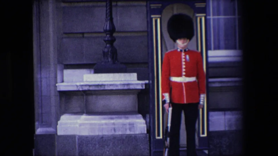 1967 London home-movie still showing a red-coated ceremonial guard beside a sentry box, with stonework, a window, and dark frame edges visible.