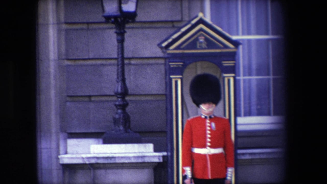 1967 London home-movie frame showing a British ceremonial guard in red uniform and black hat standing at attention beside a sentry box against a stone building facade.