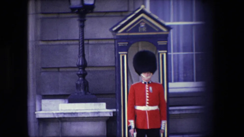 1967 London home-movie frame with a British ceremonial guard standing still in a red tunic and black hat near a sentry box, stone wall, lamp post, and window.