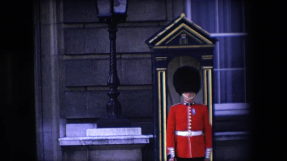 1967 London frame showing a British ceremonial guard partly centered by a sentry box, with a lamp post, stone building surface, and window visible in soft 8mm color.