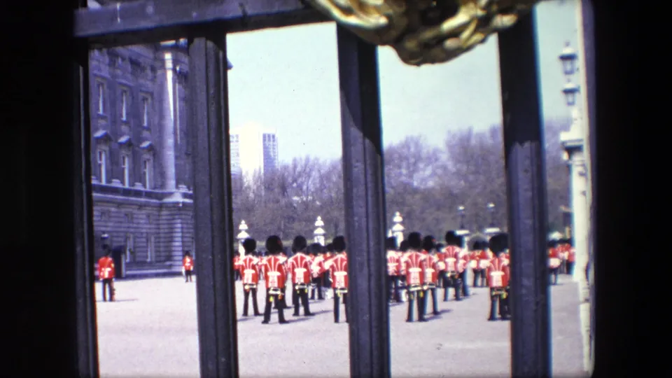 1967 London home-movie frame looking through dark gate posts toward a formation of British ceremonial guards in red uniforms, with buildings and trees in the background.