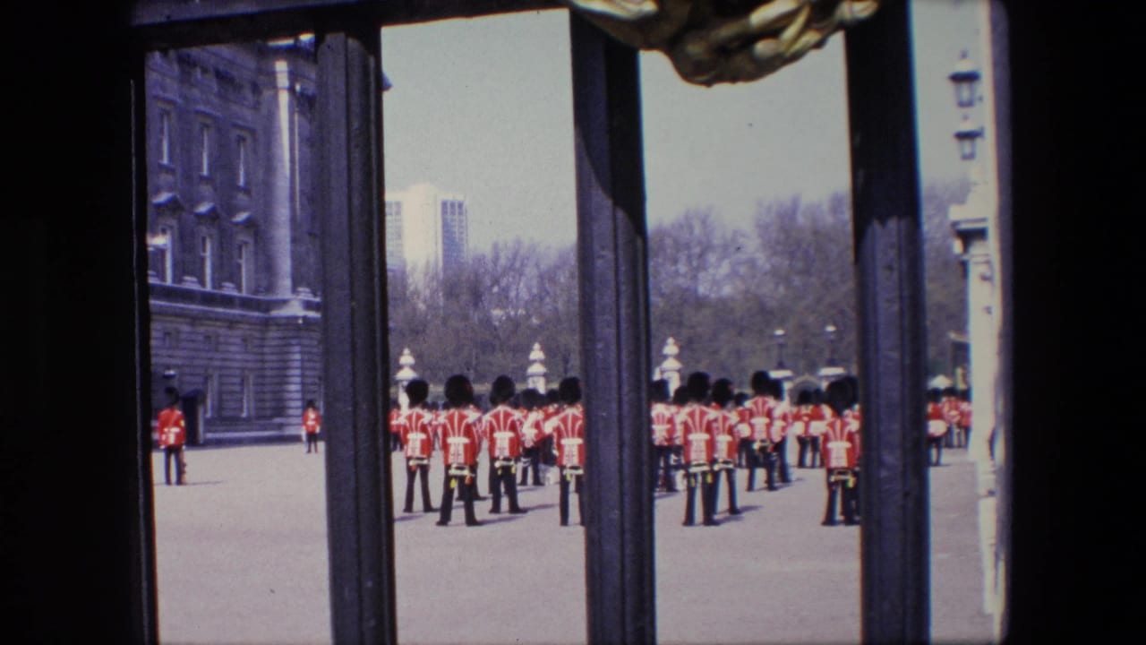 Red-uniformed figures stand in formation beyond dark gate bars on a paved London ground.