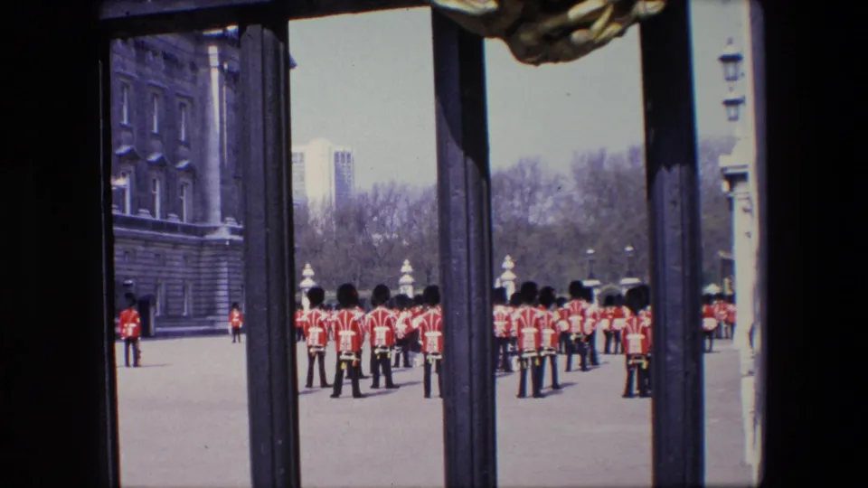 1967 London home-movie frame showing red-uniformed figures with tall black hats standing in formation beyond dark gate bars, with a stone building and trees in the background.