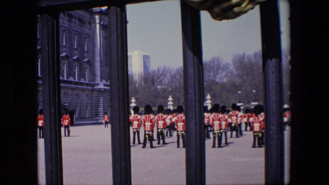 1967 London frame of red-coated uniformed figures standing in formation near a large stone facade, with the view partly blocked by dark vertical gate bars.