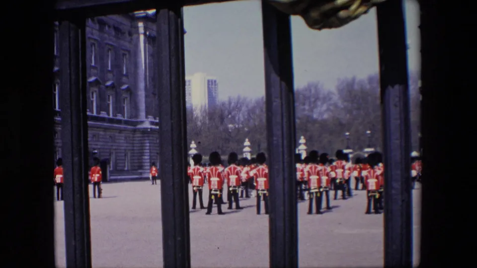 1967 London frame of red-coated uniformed figures standing in formation near a large stone facade, with the view partly blocked by dark vertical gate bars.