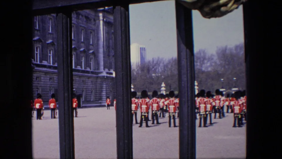 1967 London home-movie still with red-uniformed figures in tall black hats lined across a paved area, seen from behind dark railings or gates.