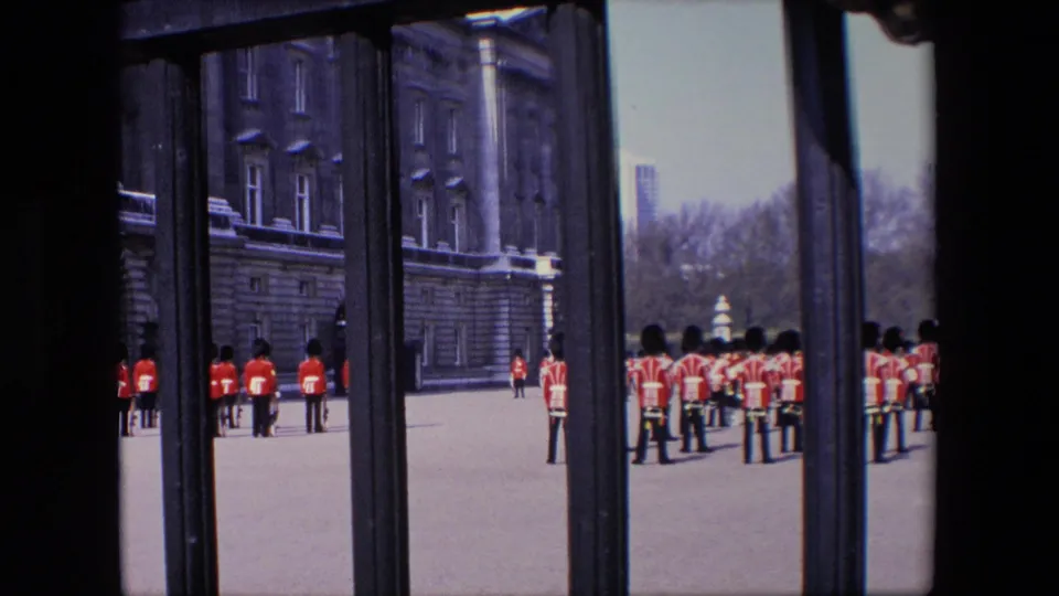 1967 London frame showing a red-uniformed ceremonial or band formation on paved grounds, viewed through dark foreground gate bars with trees and buildings beyond.