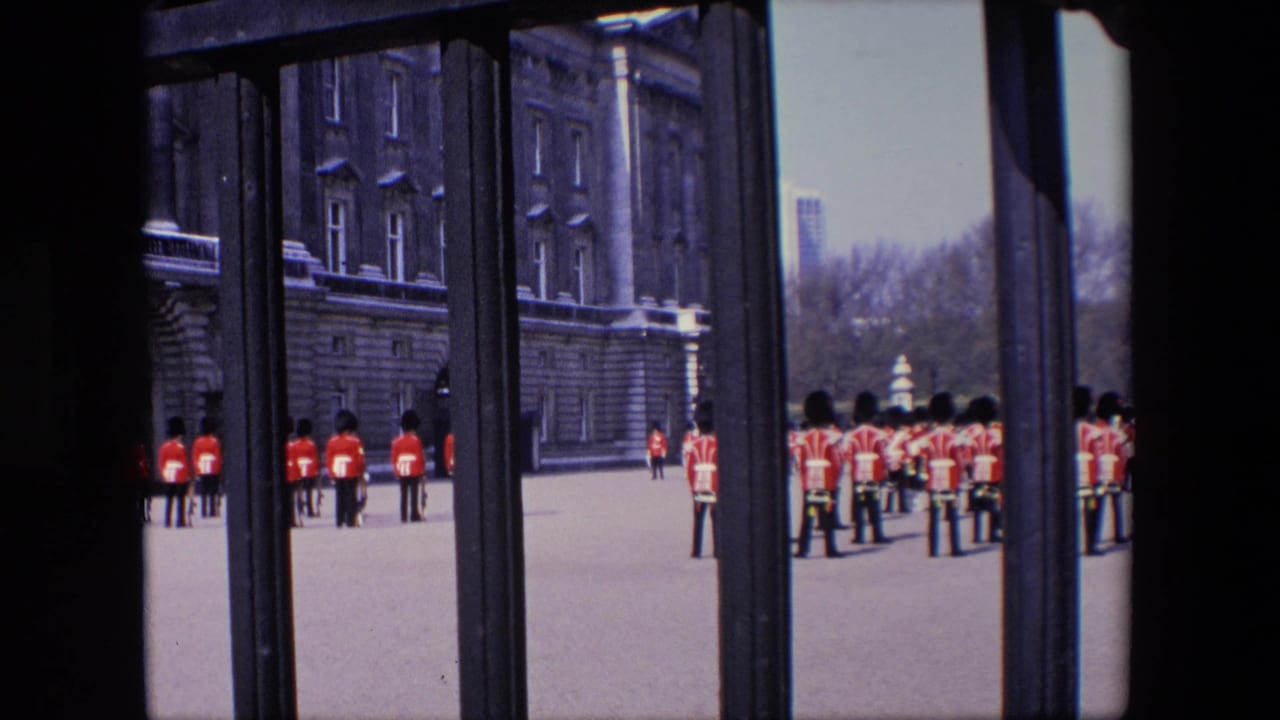 Queen's Guards in red uniforms stand in formation outside a palace, viewed through dark railings.