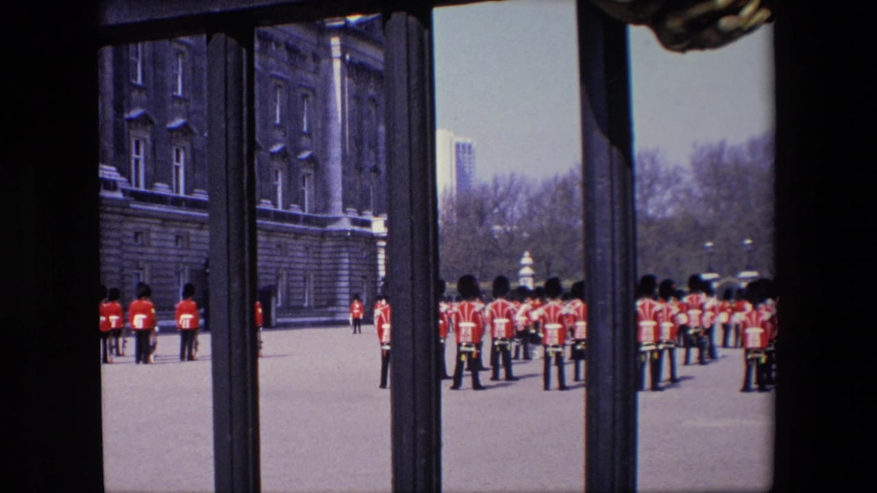 Rows of red-coated guards stand near Buckingham Palace with railings in the foreground.