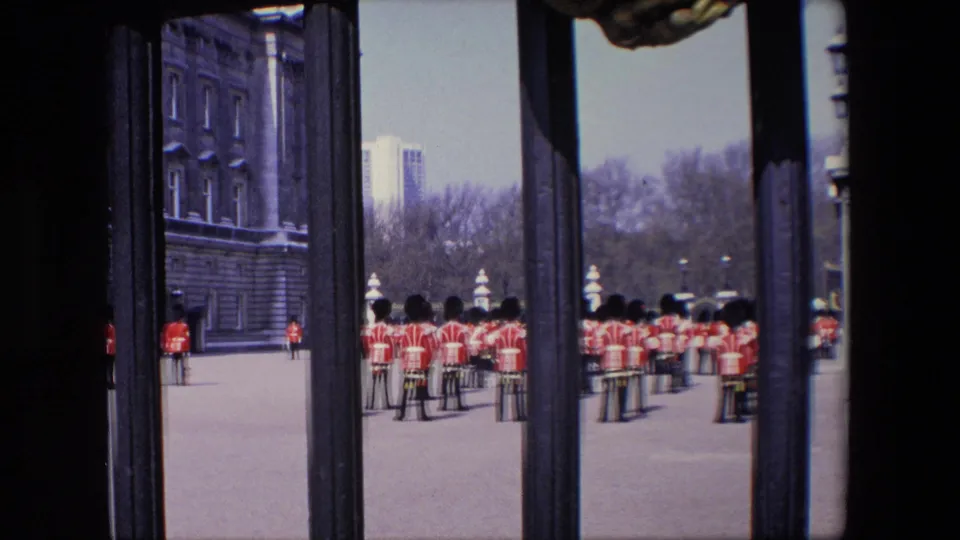 1967 London 8mm frame with Queen's Guards lined up across the paved grounds outside Buckingham Palace, seen from a partially obstructed visitor vantage point.