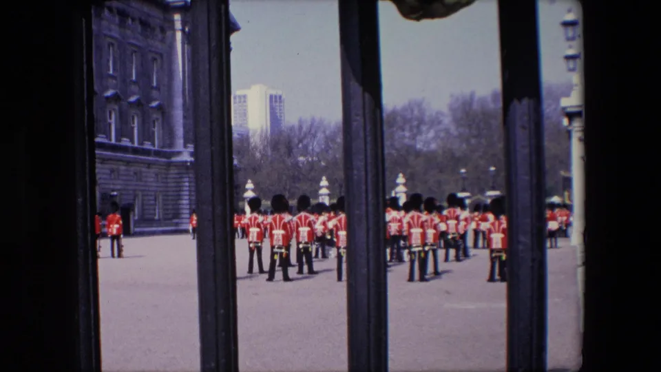 1967 London frame showing a clustered formation of Queen's Guards on the grounds outside Buckingham Palace, with the palace facade and railings shaping the composition.