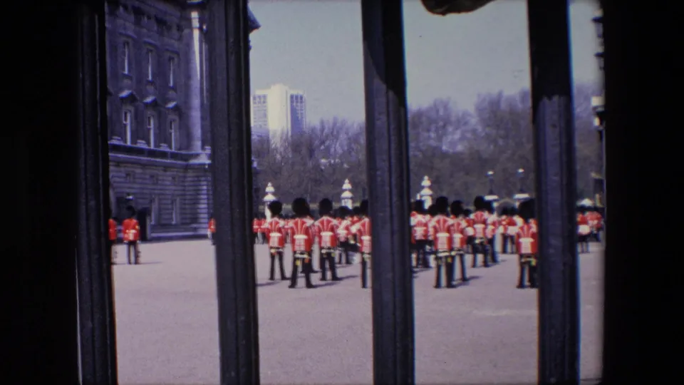 1967 London home-movie frame showing Royal Guard soldiers in red tunics and bearskin hats viewed through dark iron gate bars on a broad paved forecourt.