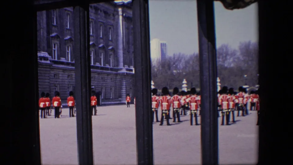 1967 London frame of Royal Guard soldiers standing at attention in red uniforms beside a large stone facade, filmed from behind iron gates.