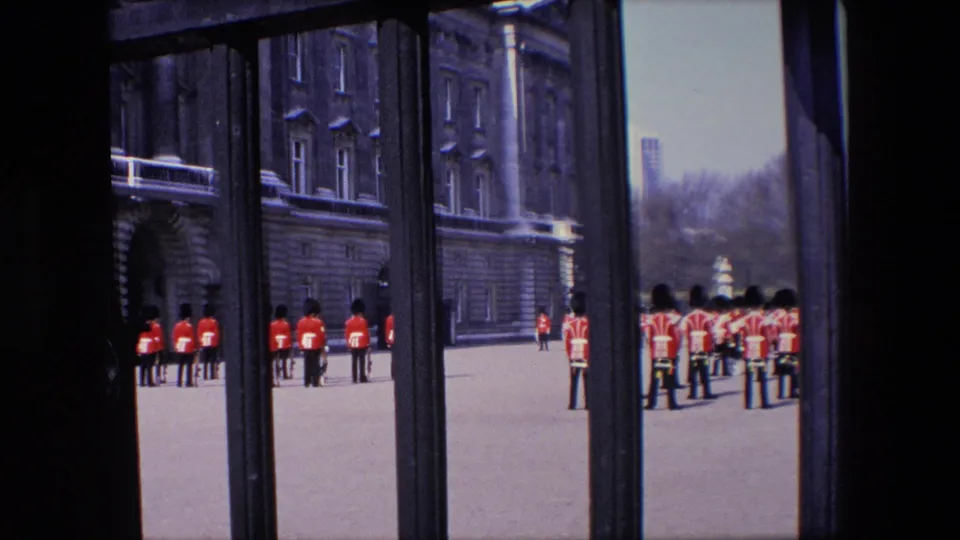 1967 London home-movie view of Royal Guard members in red tunics standing near a stone building, with the scene partly framed by dark gate bars.