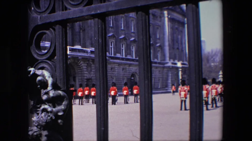 1967 London frame showing Royal Guard soldiers in formation behind ornate ironwork, with a stone building and open forecourt visible beyond the gate.