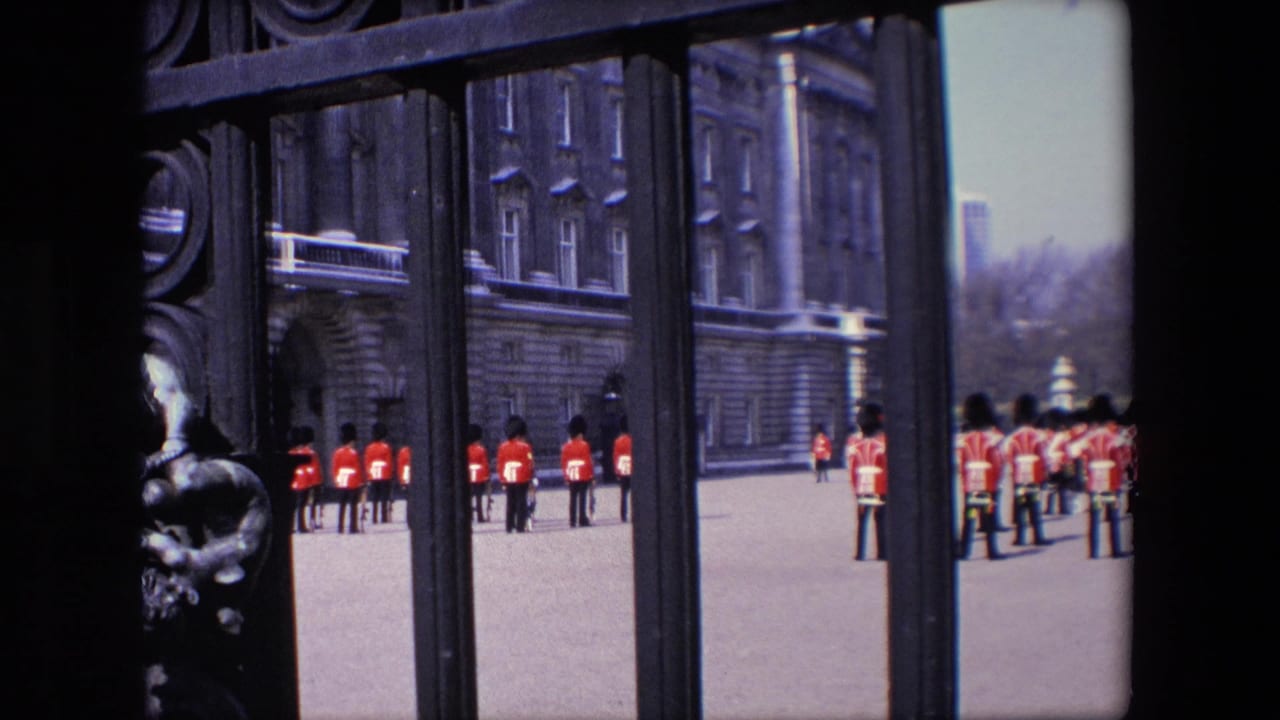 1967 London home-movie frame showing royal guards in red uniforms and tall dark hats standing near a palace gate with stone buildings behind them.