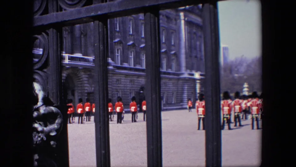1967 London home-movie frame showing royal guards in red uniforms and tall dark hats standing near a palace gate with stone buildings behind them.