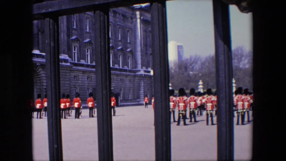 1967 London frame with multiple royal guards in red uniforms visible through gate bars, with palace architecture and trees in the background.