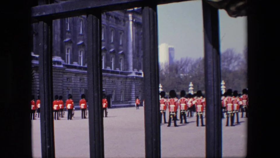 1967 London home-movie view of royal guards at a palace forecourt, partly obstructed by dark gate bars and a stone facade at left.