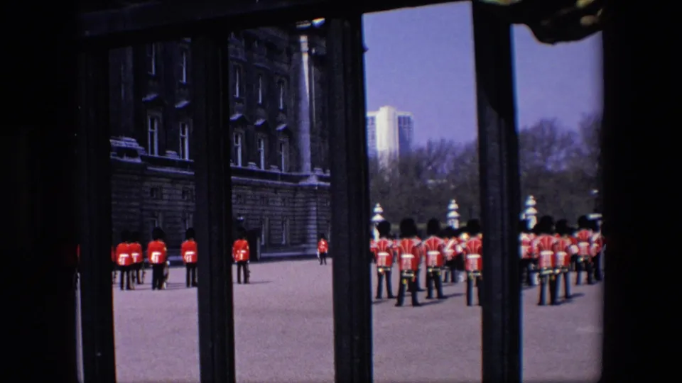 1967 London frame showing rows of red-uniformed royal guards beyond an iron gate, with palace stonework, trees, and a distant building visible.