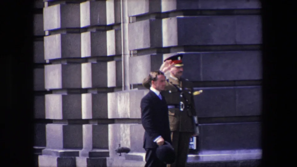 1967 London frame with a uniformed officer raising a salute beside a suited man holding a bowler-style hat near a stone facade.