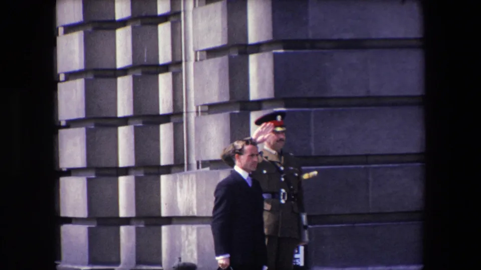 1967 London home-movie frame showing a suited man passing in front of a uniformed officer positioned against a stone exterior.