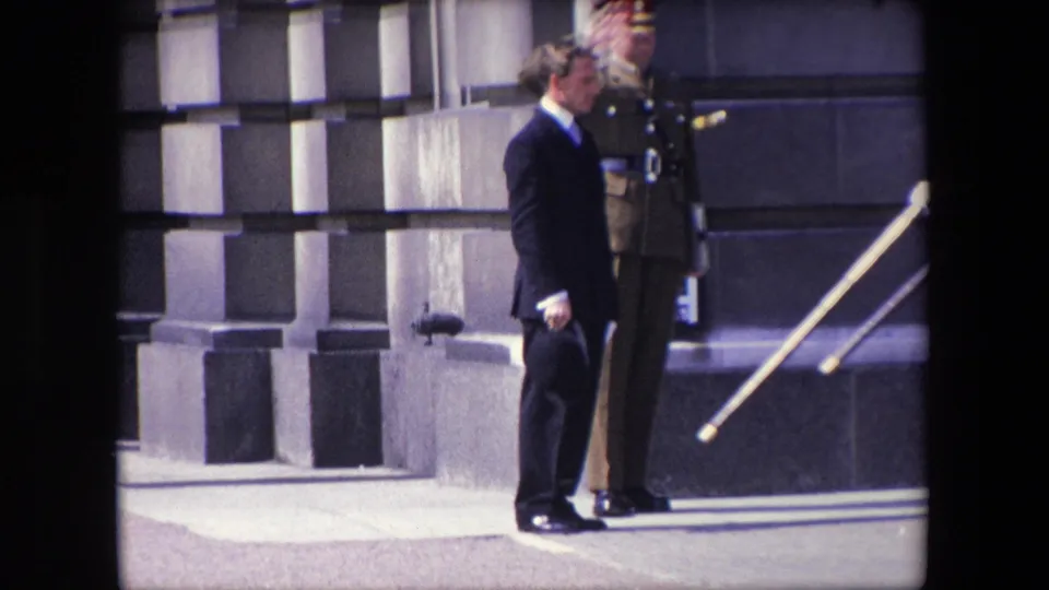 1967 London frame showing a suited man holding a dark hat near a uniformed officer, with steps or railings visible at the edge of the formal exterior.