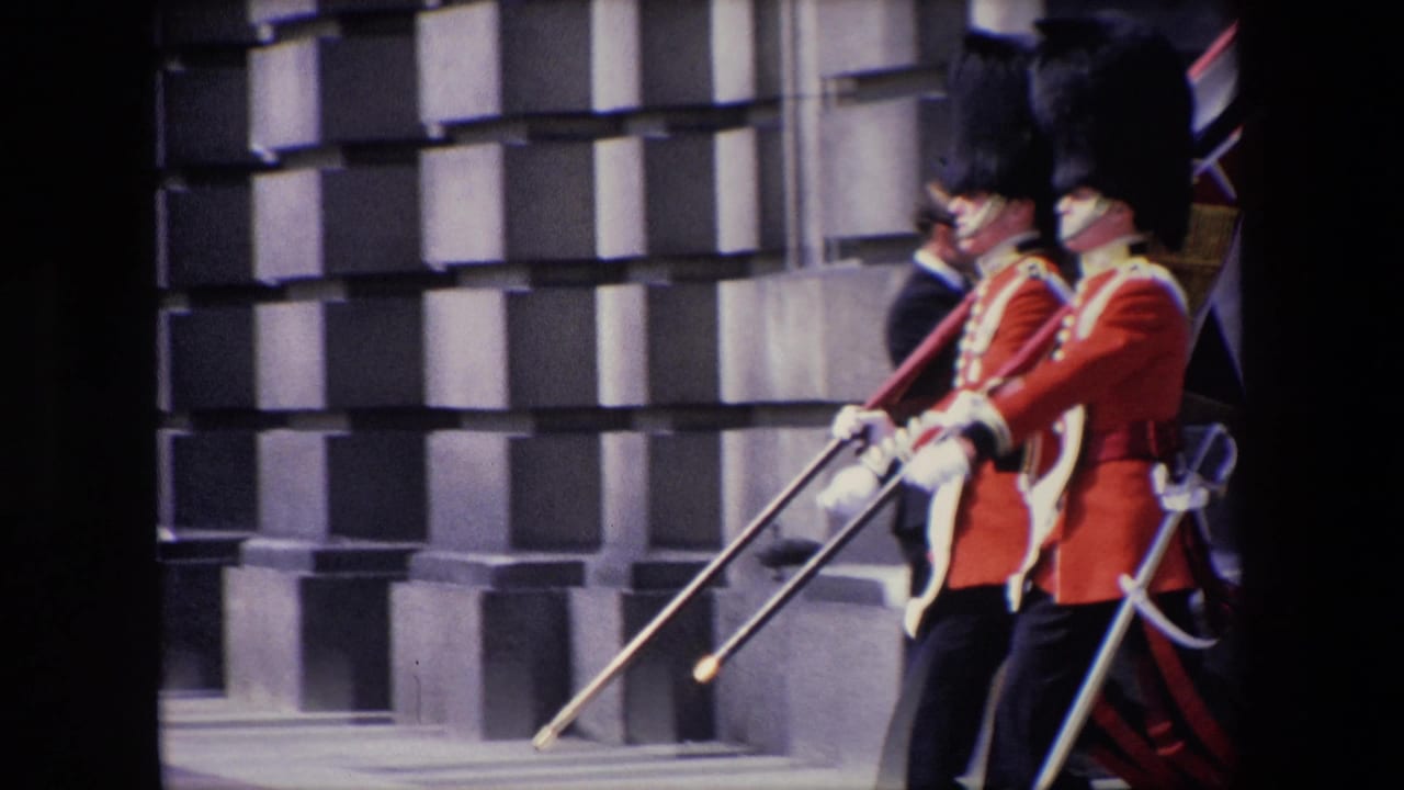 Two guards in red ceremonial uniforms march beside a stone building.