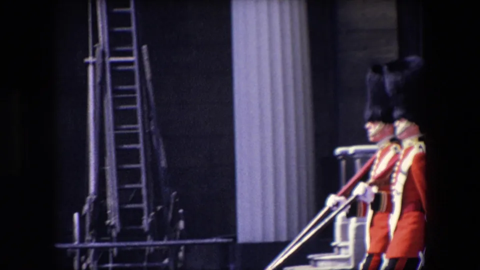 Two guards in red ceremonial dress appear near a columned building and vertical ladder-like structure during the London guard ceremony.