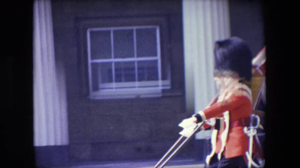 A single guard in red ceremonial uniform and tall black hat is partly framed by a window, column, and dark film border in the 1967 London home-movie footage.