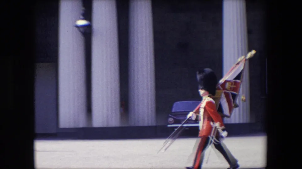 A ceremonial guard in red uniform carries a flag while marching across an open area near building columns and a parked car in London.