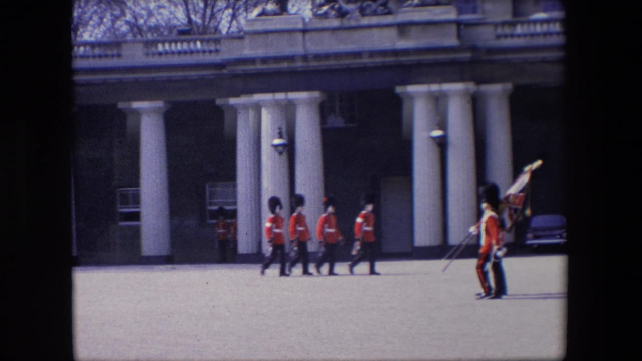British guards in red uniforms march across an open ground near white columns.