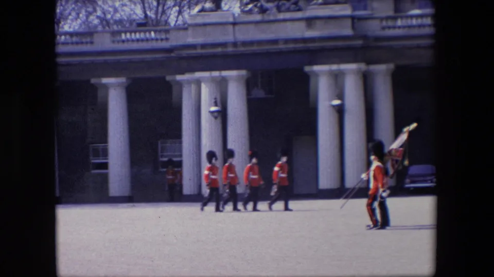 In 1967 London, two British guards in red uniforms and bearskin-style hats carry flags while marching across an open parade ground near a colonnaded building.