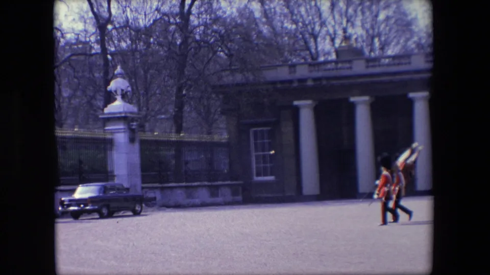Two flag-bearing British guards march across the open ground while a stone bust, railings, parked dark car, and columned building remain visible behind them.