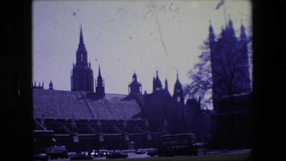 London landmark architecture appears in silhouette above a roadway with period cars and a red double-decker bus, filmed in high-contrast 8mm color.