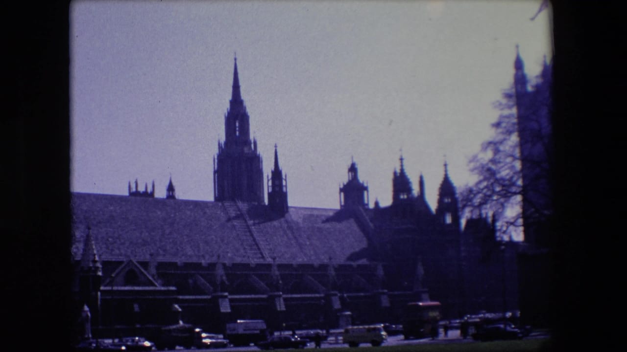 Traffic passes in front of a large spired building seen from across the road.
