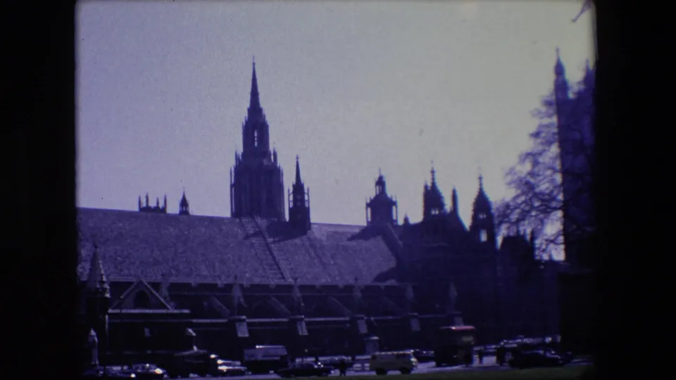 1967 London home-movie frame showing cars and larger vehicles moving along a road before a large Gothic-style building with a tall central spire.