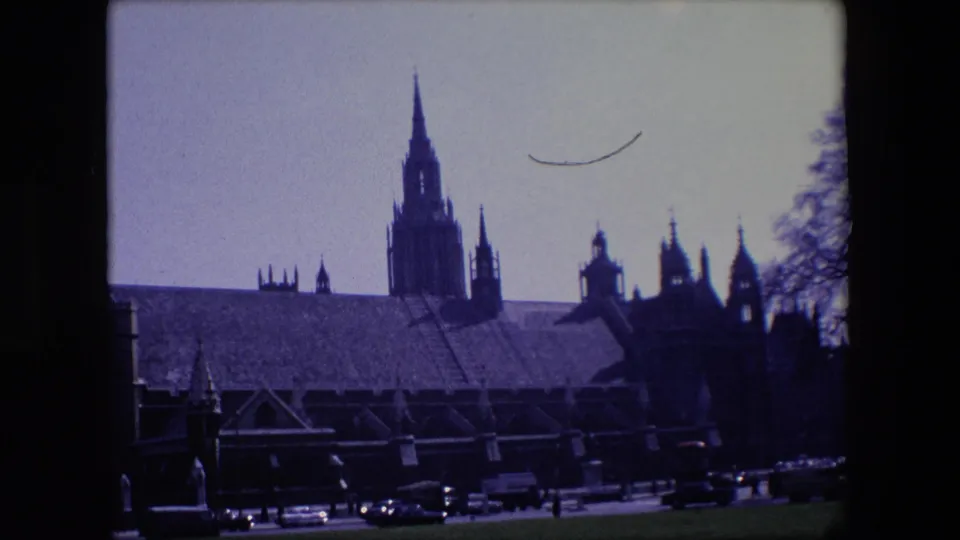 1967 London street view with a Gothic-style building, silhouetted towers, cars, and a larger vehicle along the foreground road.