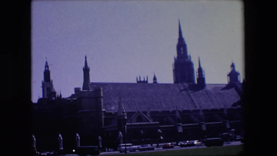 1967 London home-movie frame showing the roofline and tall spire of a large building above cars moving through the lower edge of the shot.