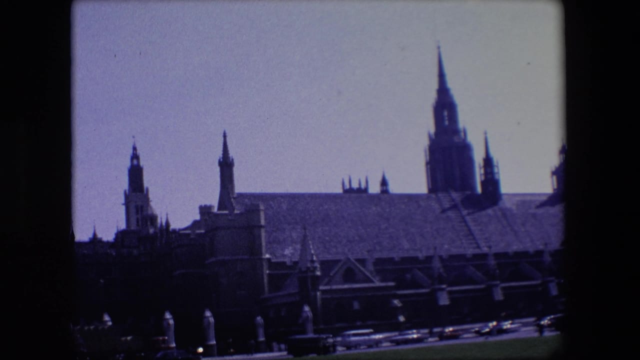 1967 London home-movie frame showing a large Gothic building with towers and buttresses behind passing road traffic.
