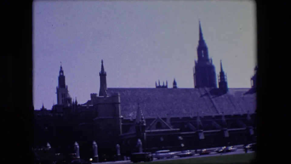1967 London home-movie frame showing a large Gothic building with towers and buttresses behind passing road traffic.