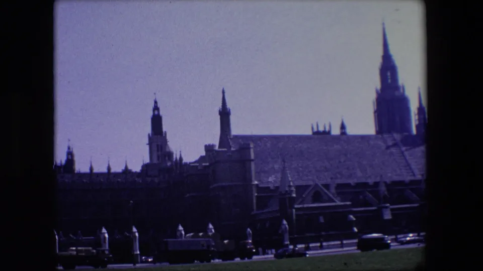 1967 London frame with buses, vans, and cars moving along the road below a Gothic landmark building and tall spire.