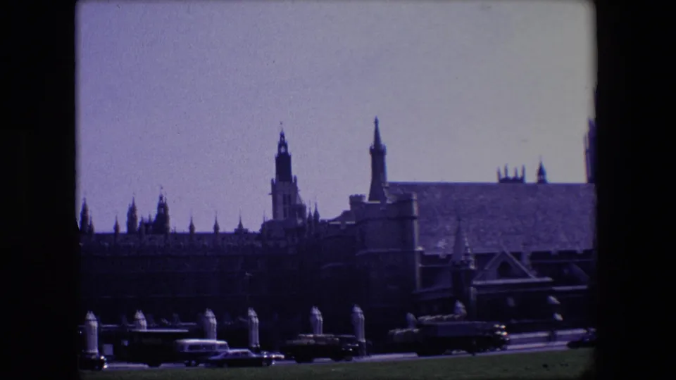 1967 London street view with heavy vehicle traffic beneath Gothic towers, buttresses, and rooflines in a handheld 8mm frame.