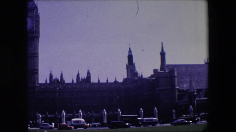 1967 London frame showing a prominent clock tower, Gothic roofline, roadside posts, buses, and cars in front of historic architecture.