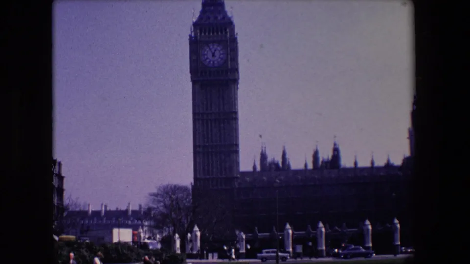 1967 London home-movie view of Big Ben and the Palace of Westminster with vehicles and trees in the foreground.