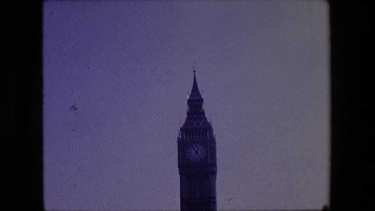 1967 London home-movie frame showing the upper Elizabeth Tower clock face and spire against a pale sky, with soft focus and visible 8mm frame edges.