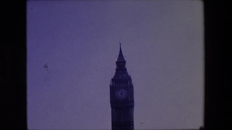 1967 London home-movie frame showing the upper Elizabeth Tower clock face and spire against a pale sky, with soft focus and visible 8mm frame edges.