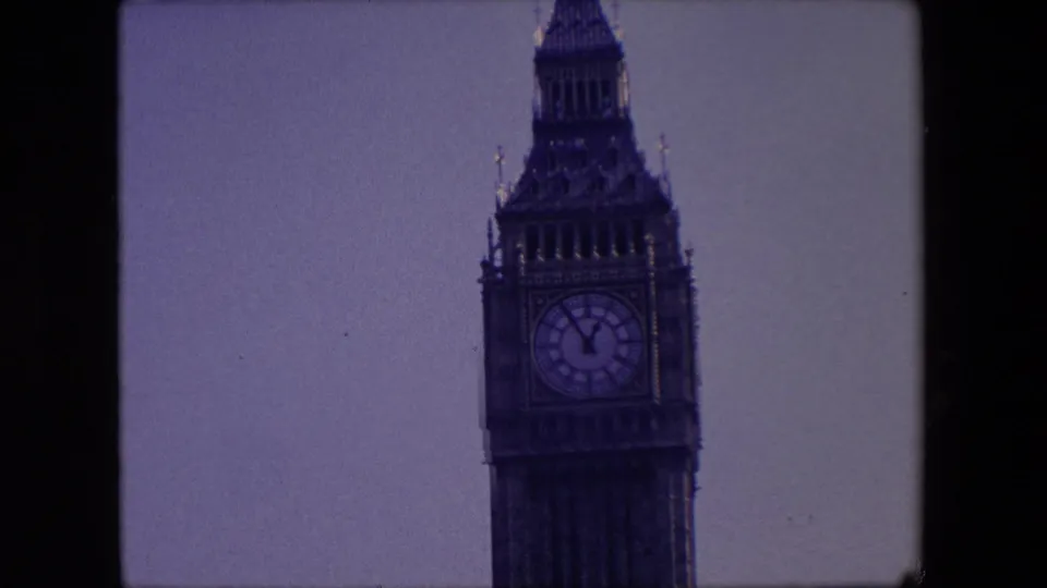 Close 8mm home-movie view of the Elizabeth Tower clock face and spire in London, with the tower filling more of the frame and pale sky behind it.