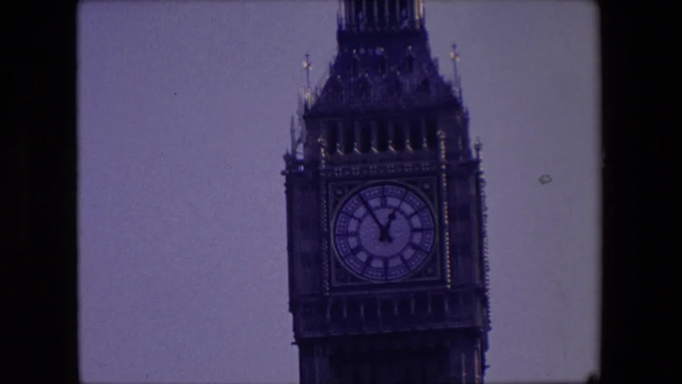 1967 London home-movie frame showing the Elizabeth Tower clock face from a distance, with pale sky, soft focus, and black film borders.