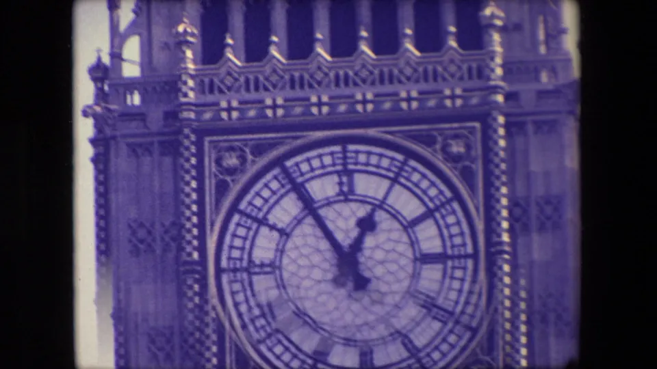 1967 London home-movie close-up of the Westminster clock face, with Roman numerals, dark hands, decorative trim, and soft blue-purple film color.